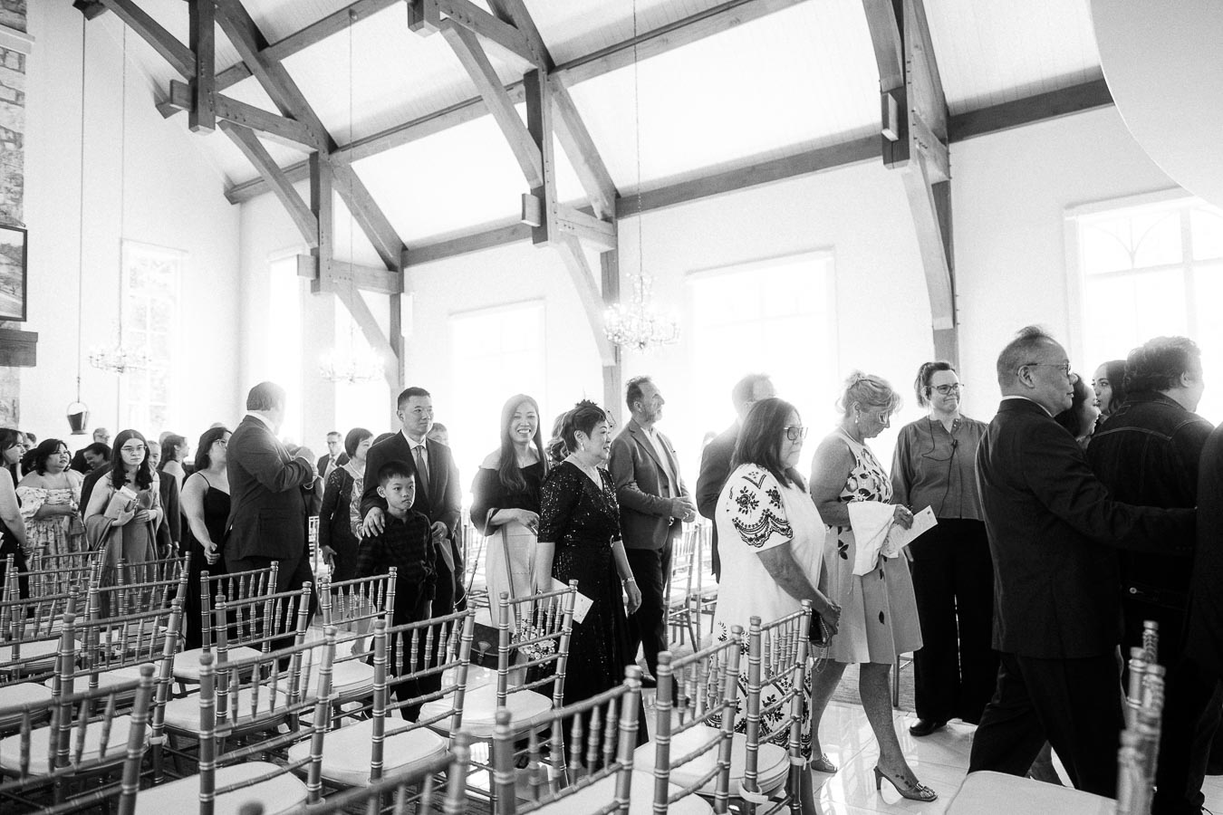 Wedding guests line up between chiavari chairs inside a bright timber-beamed chapel before the ceremony.