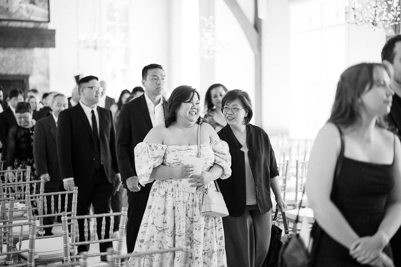 Wedding guests walk between ceremony chairs in a bright room, with two women smiling together in the aisle.
