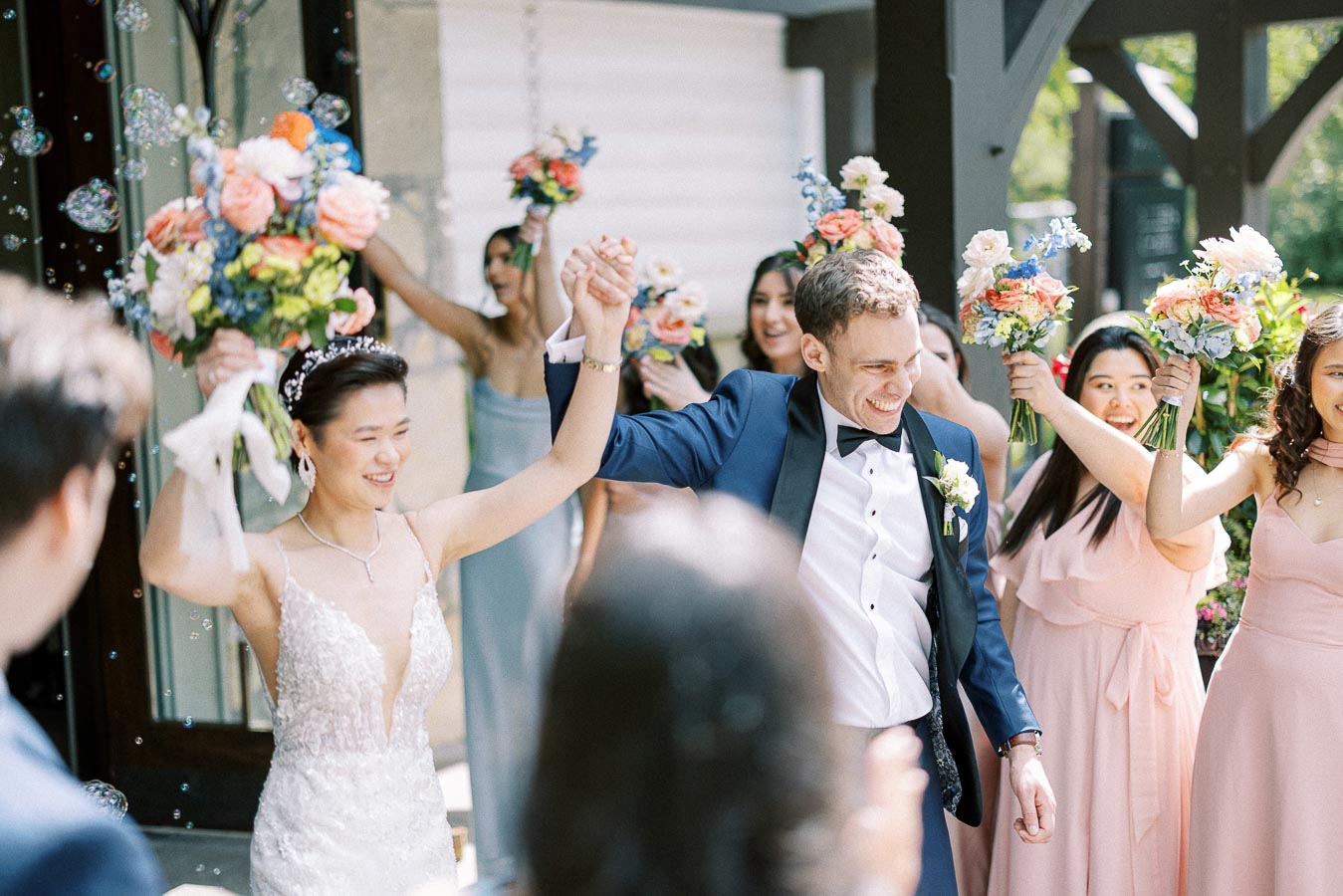 Miffy and Rawle smile with raised hands as bridesmaids hold bouquets and bubbles float around them.