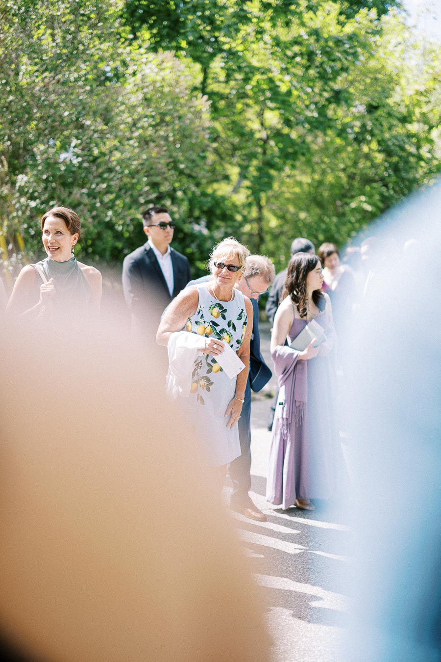 Wedding guests in formal attire gather outdoors in bright sunlight beside leafy trees at Ancaster Mill.