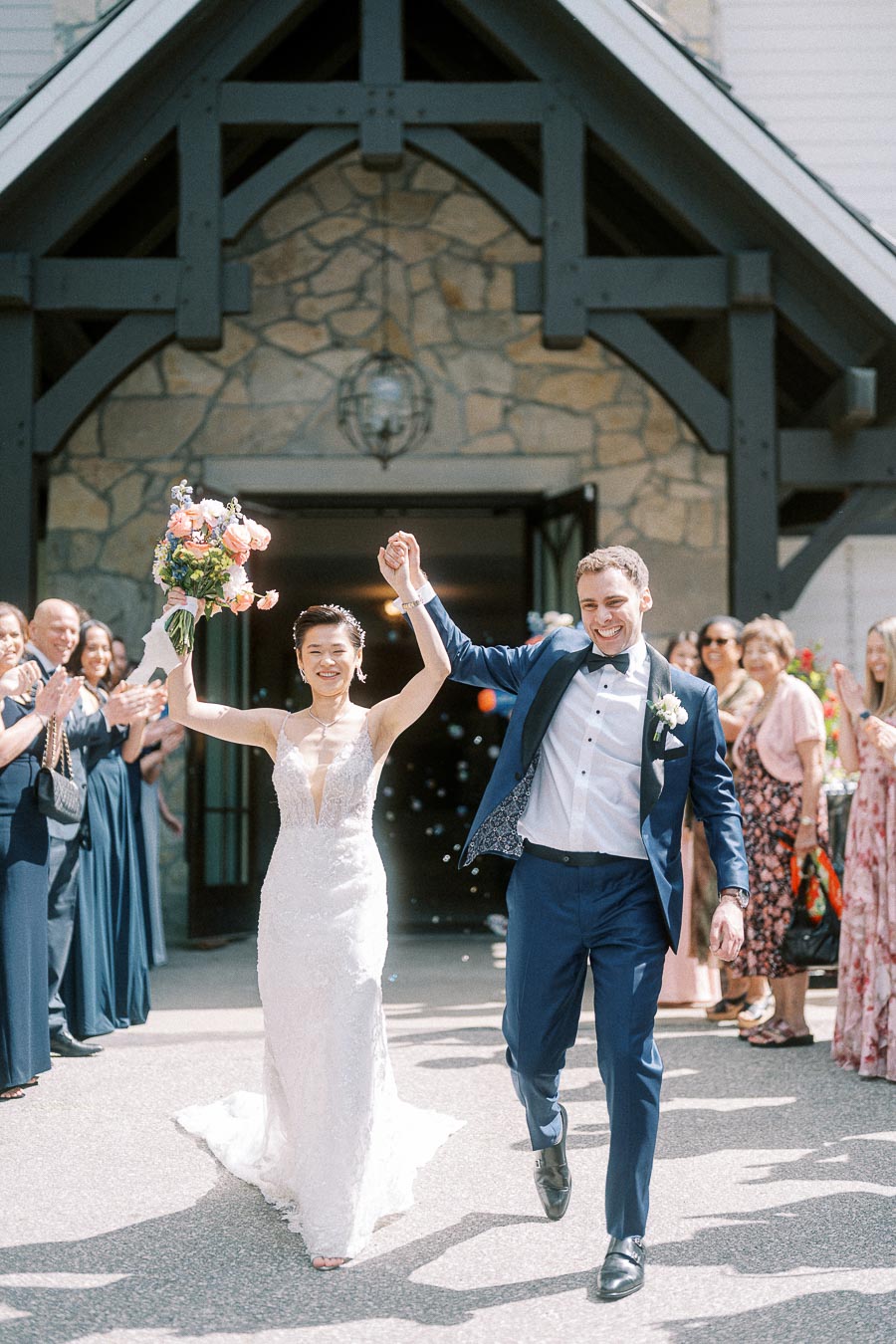 Miffy and Rawle walk hand in hand past cheering guests outside the stone chapel at Ancaster Mill.
