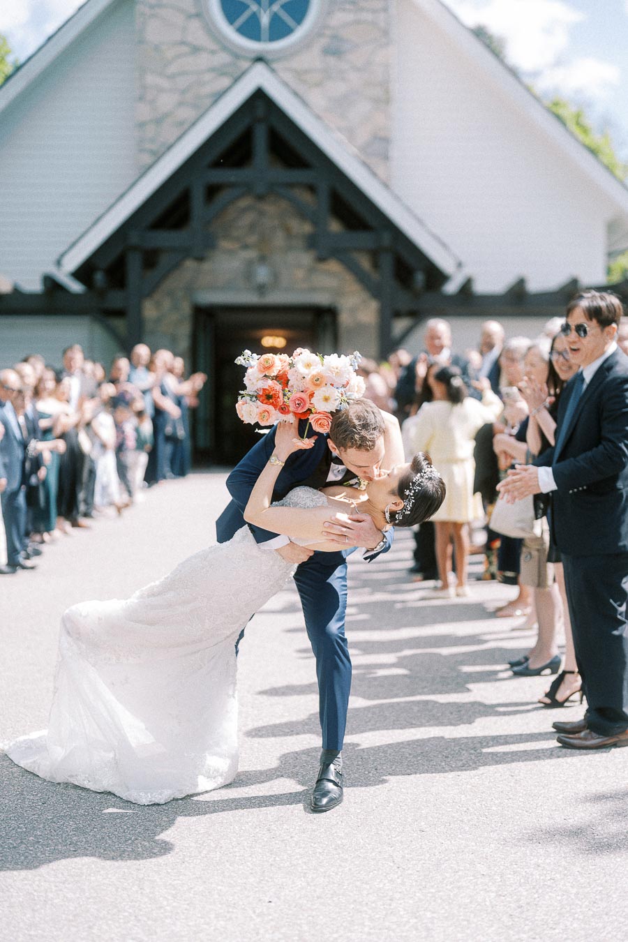 Newlyweds kiss in a dramatic dip outside a chapel as guests clap on both sides of the walkway.