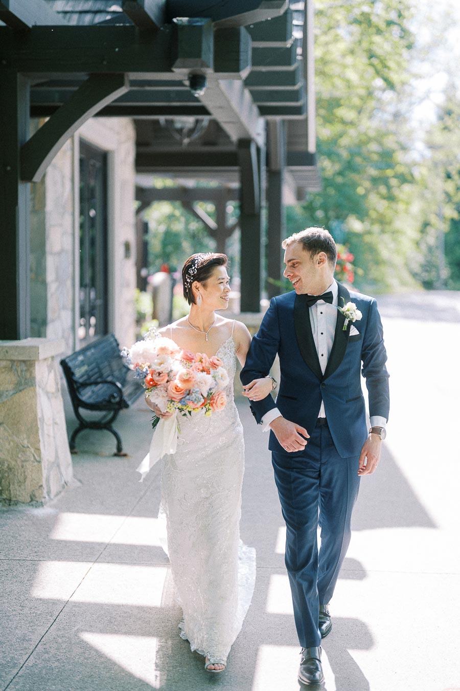 Miffy and Rawle walk arm in arm outside Ancaster Mill, smiling in wedding attire with a pastel bouquet.