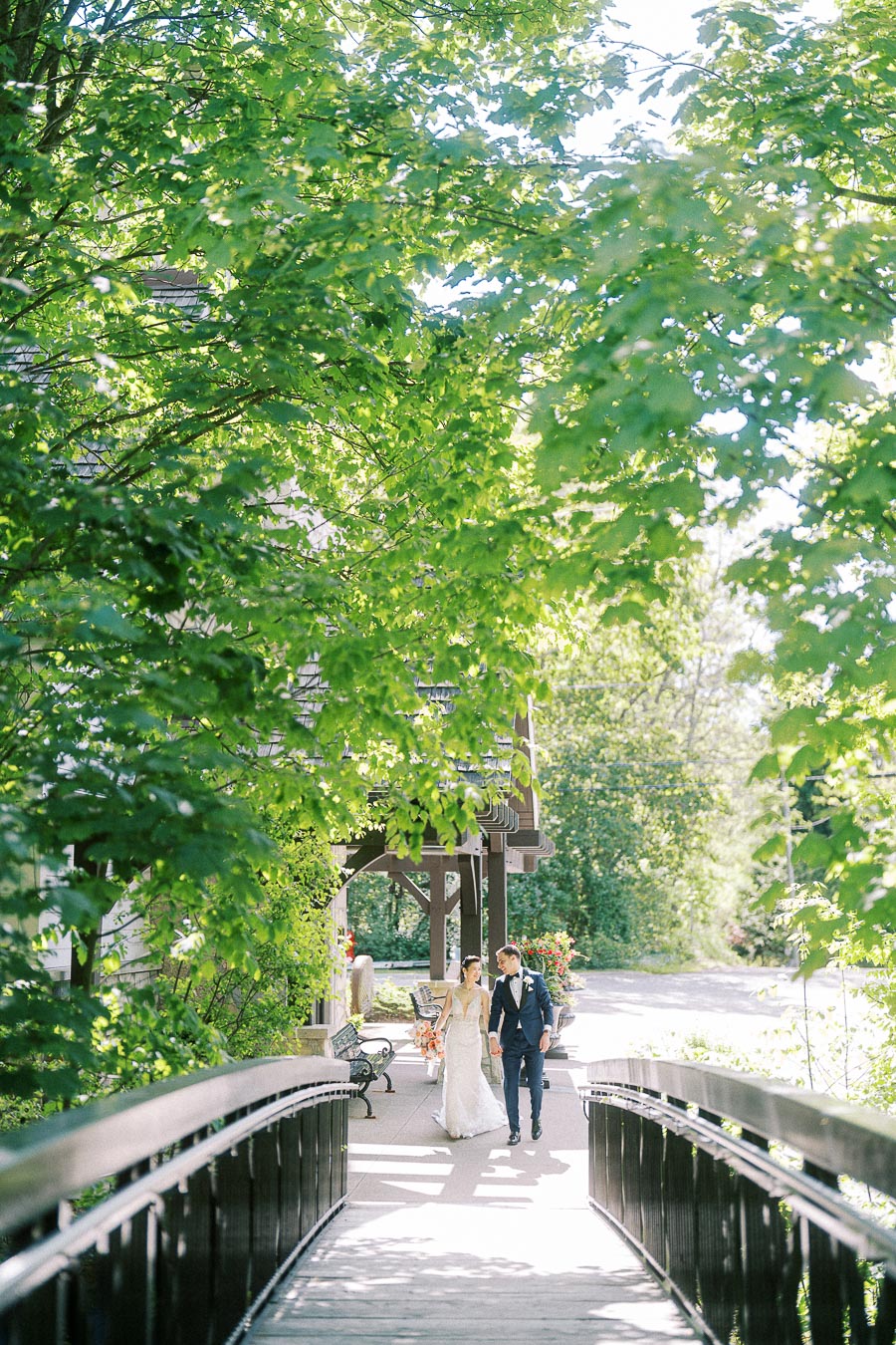 Bride and groom walk hand in hand past a wooden bridge beneath bright green trees at Ancaster Mill.