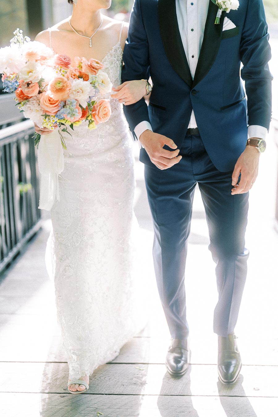 Bride with pastel bouquet walks arm in arm with groom in a navy tuxedo on a sunlit wedding walkway.
