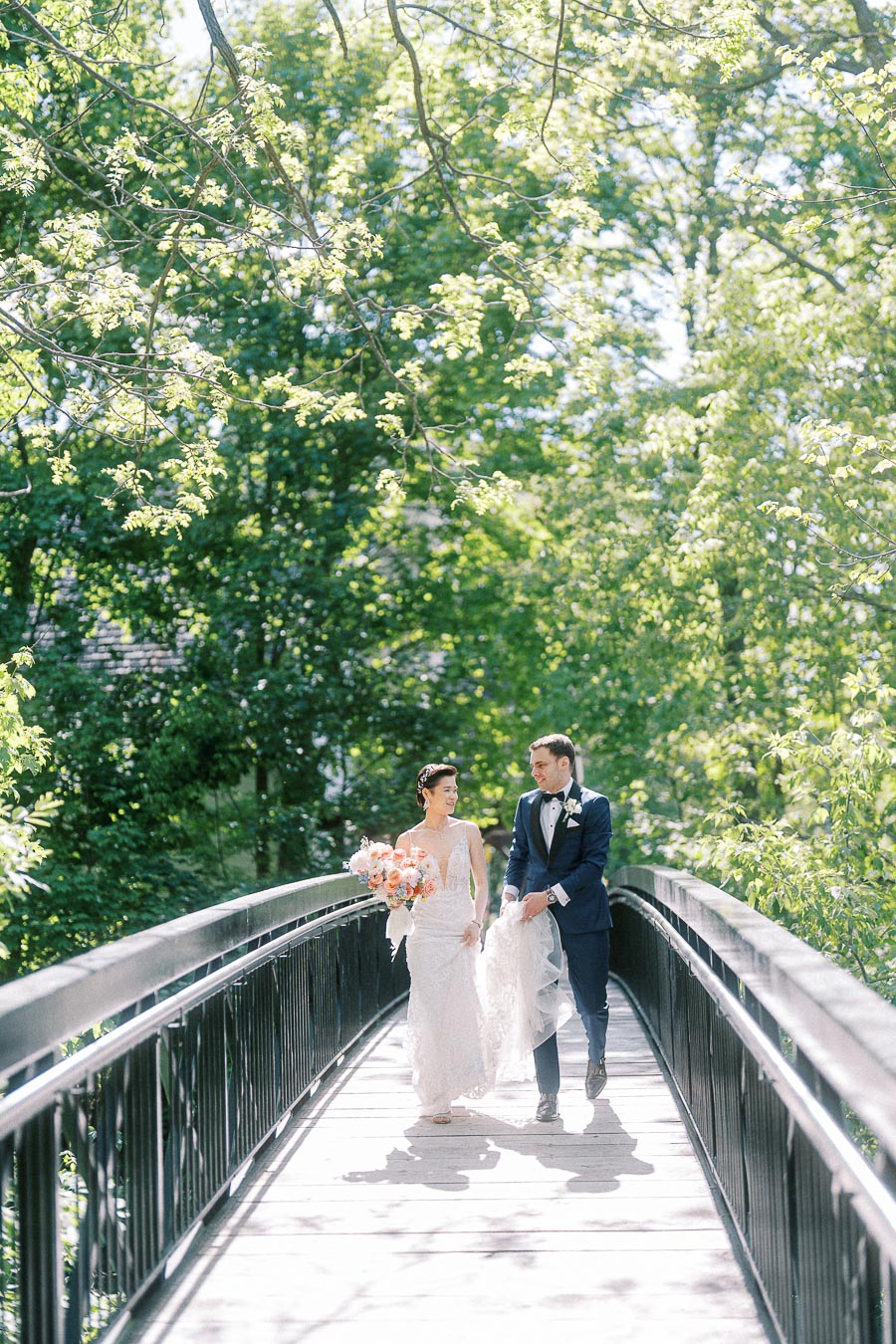 Bride and groom walk together on a sunlit bridge surrounded by green trees at Ancaster Mill.