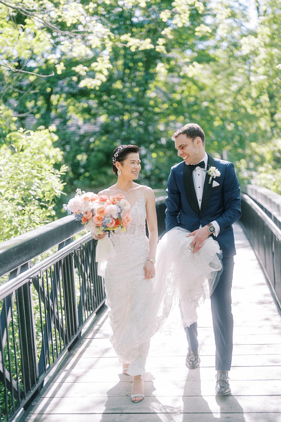 Miffy and Rawle walk together on a sunlit bridge, smiling in wedding attire with a pastel bouquet.