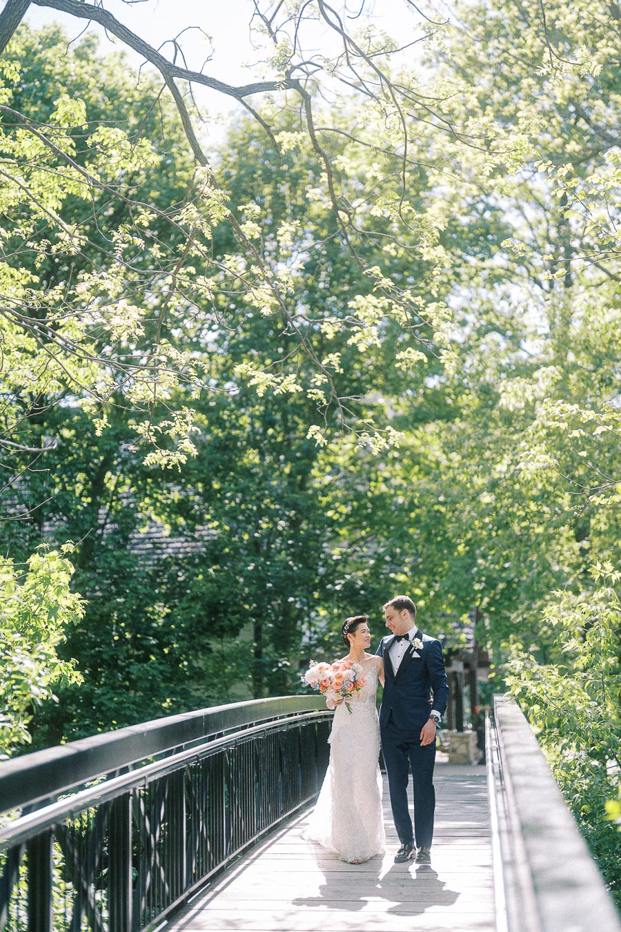 Bride and groom walk arm in arm on a sunlit bridge surrounded by green trees at Ancaster Mill.