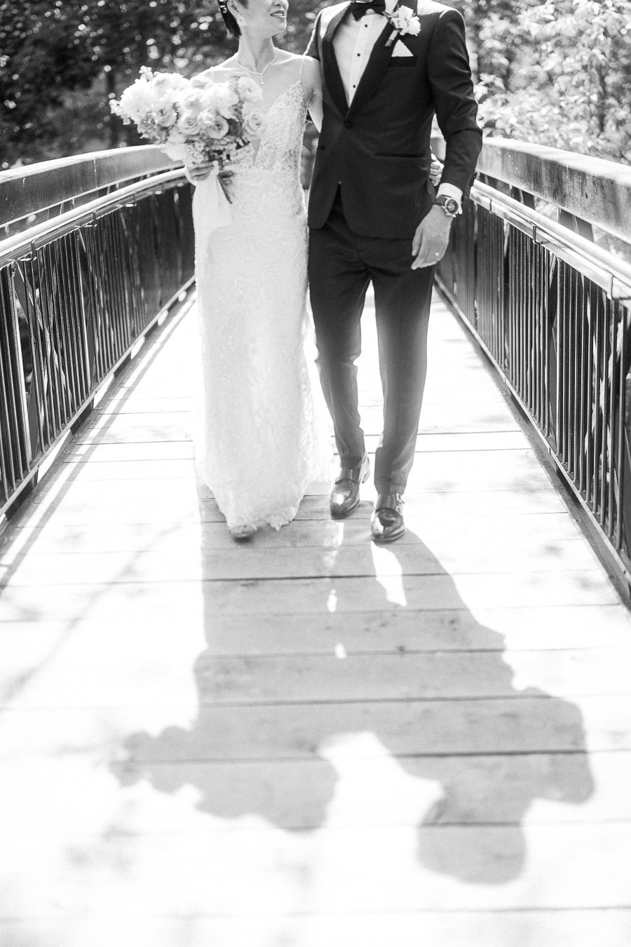 Bride and groom walk arm in arm on a sunlit bridge, with the bride holding a bouquet and their shadows on the boards.