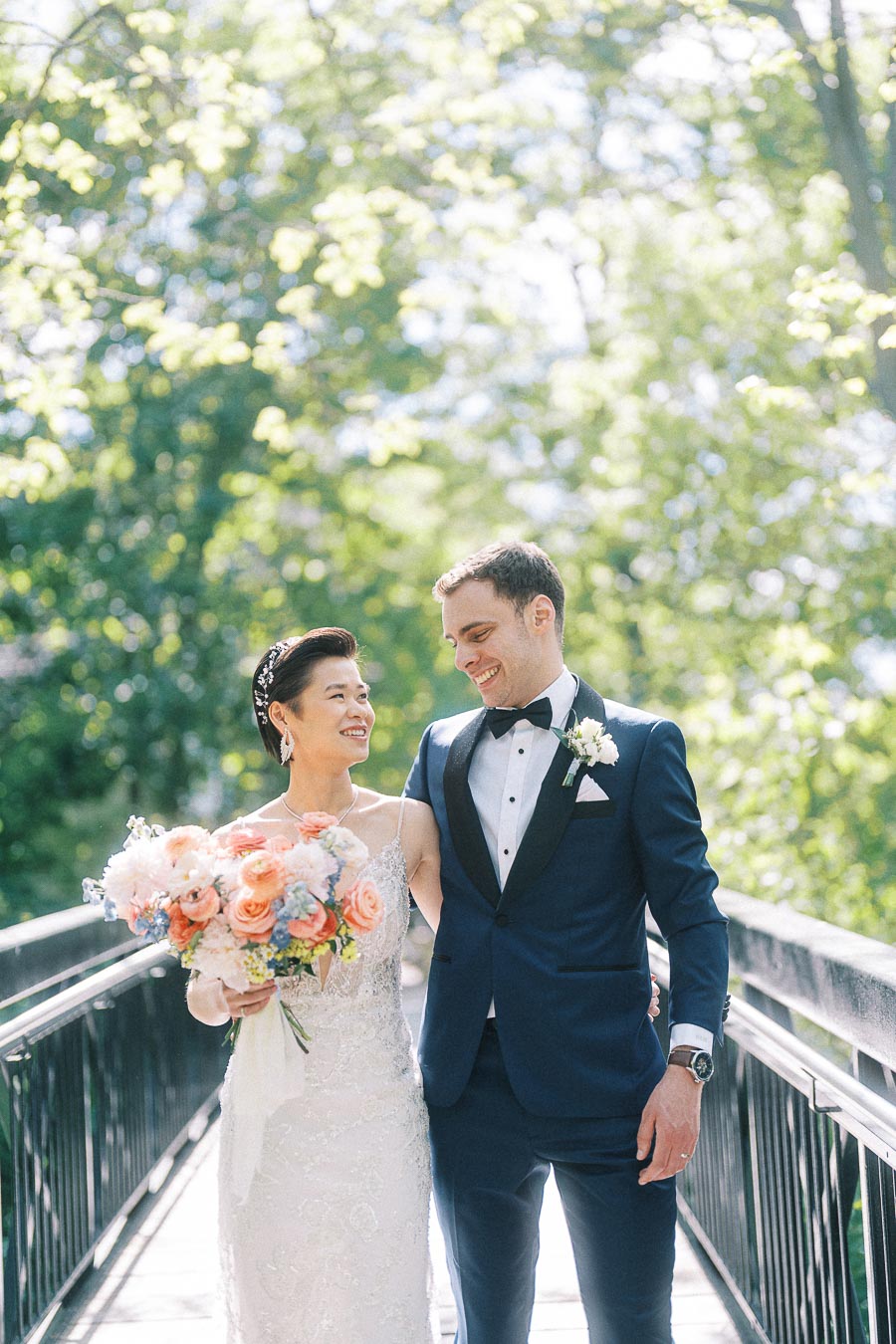 Miffy and Rawle walk arm in arm on a sunlit bridge, holding a peach and white bridal bouquet.