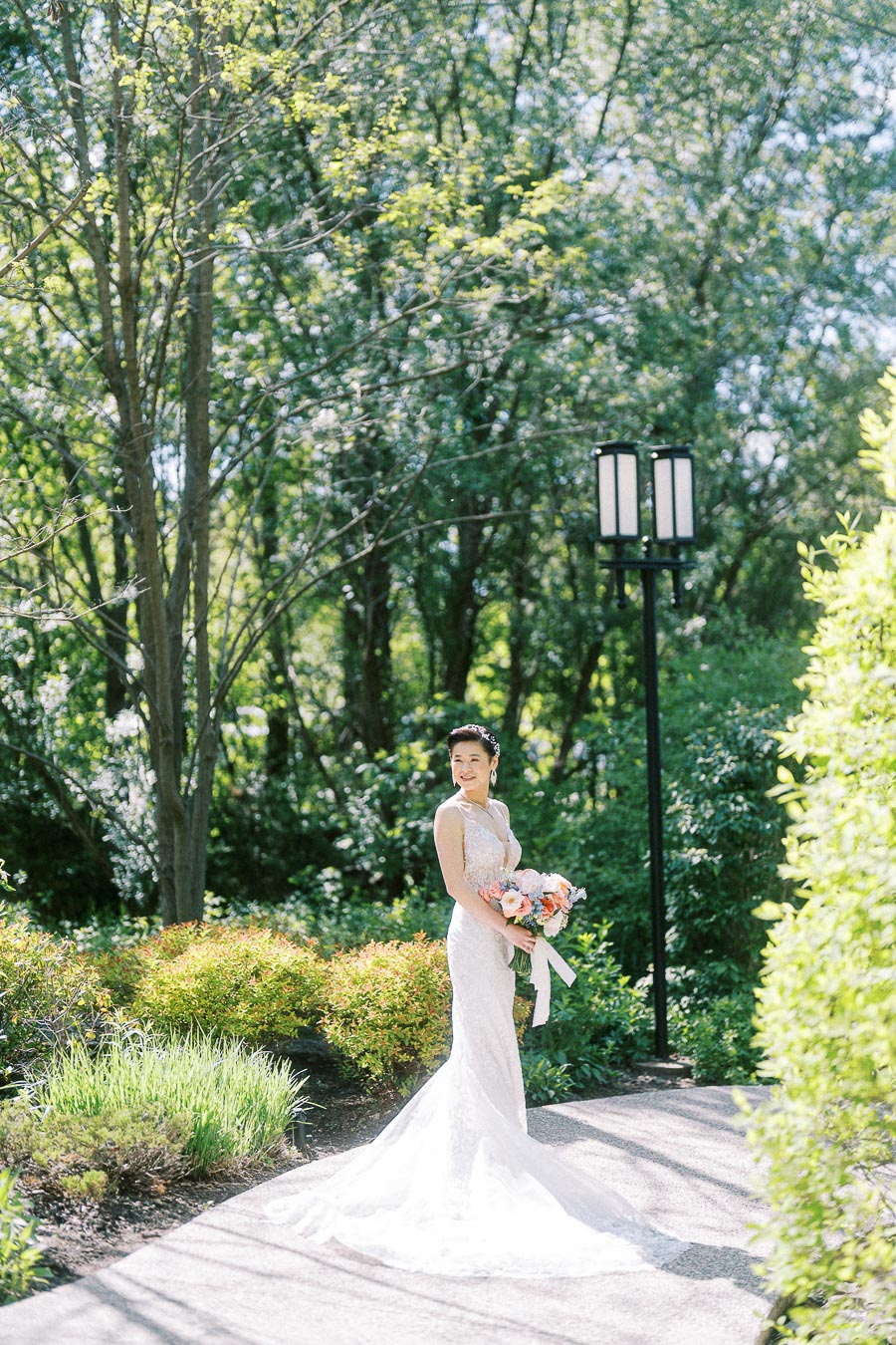 Bride in a lace wedding gown holding a peach bouquet on a sunlit garden path at Ancaster Mill.