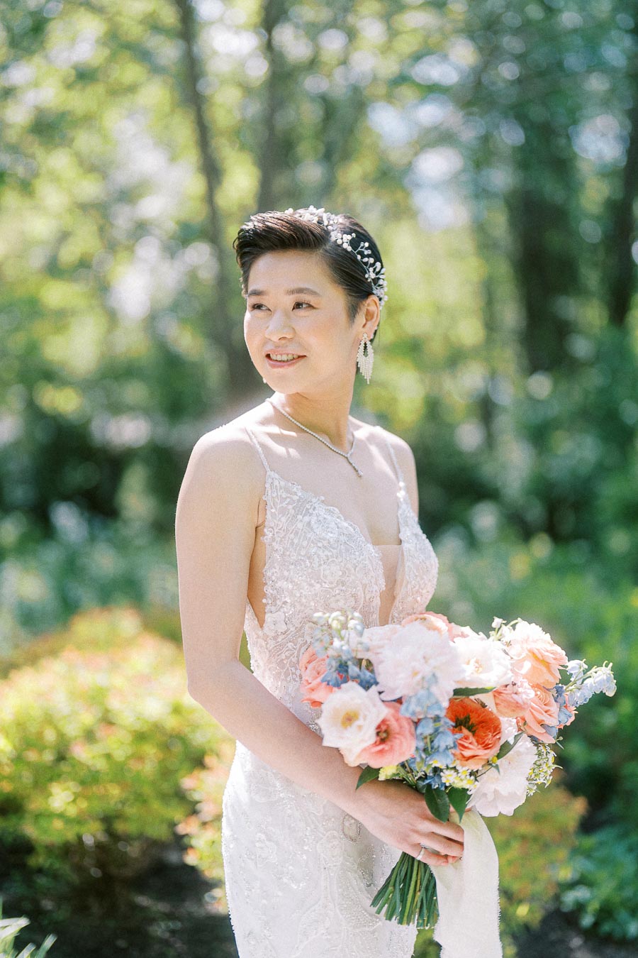 Bride in a lace wedding dress holding a pastel bouquet outdoors among sunlit trees and greenery.