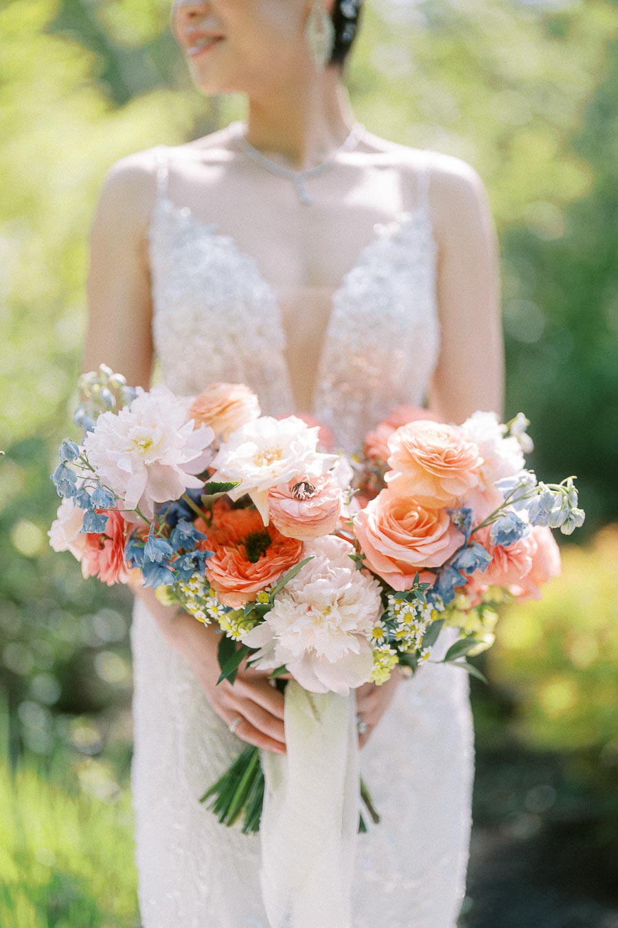 Bride in a beaded gown holding a pastel bouquet of peach roses, white flowers, and blue blooms outdoors