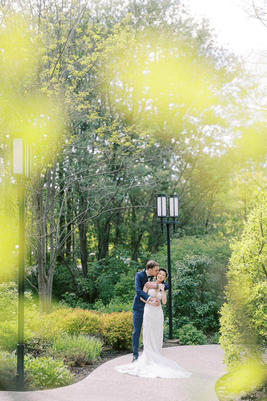 Bride in a white gown and groom in a blue suit embrace on a garden path surrounded by trees and greenery.