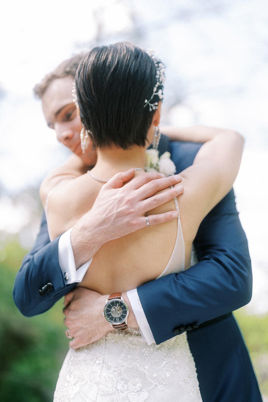 Groom in a navy suit embraces the bride from behind, his ring and watch visible against her beaded wedding dress.