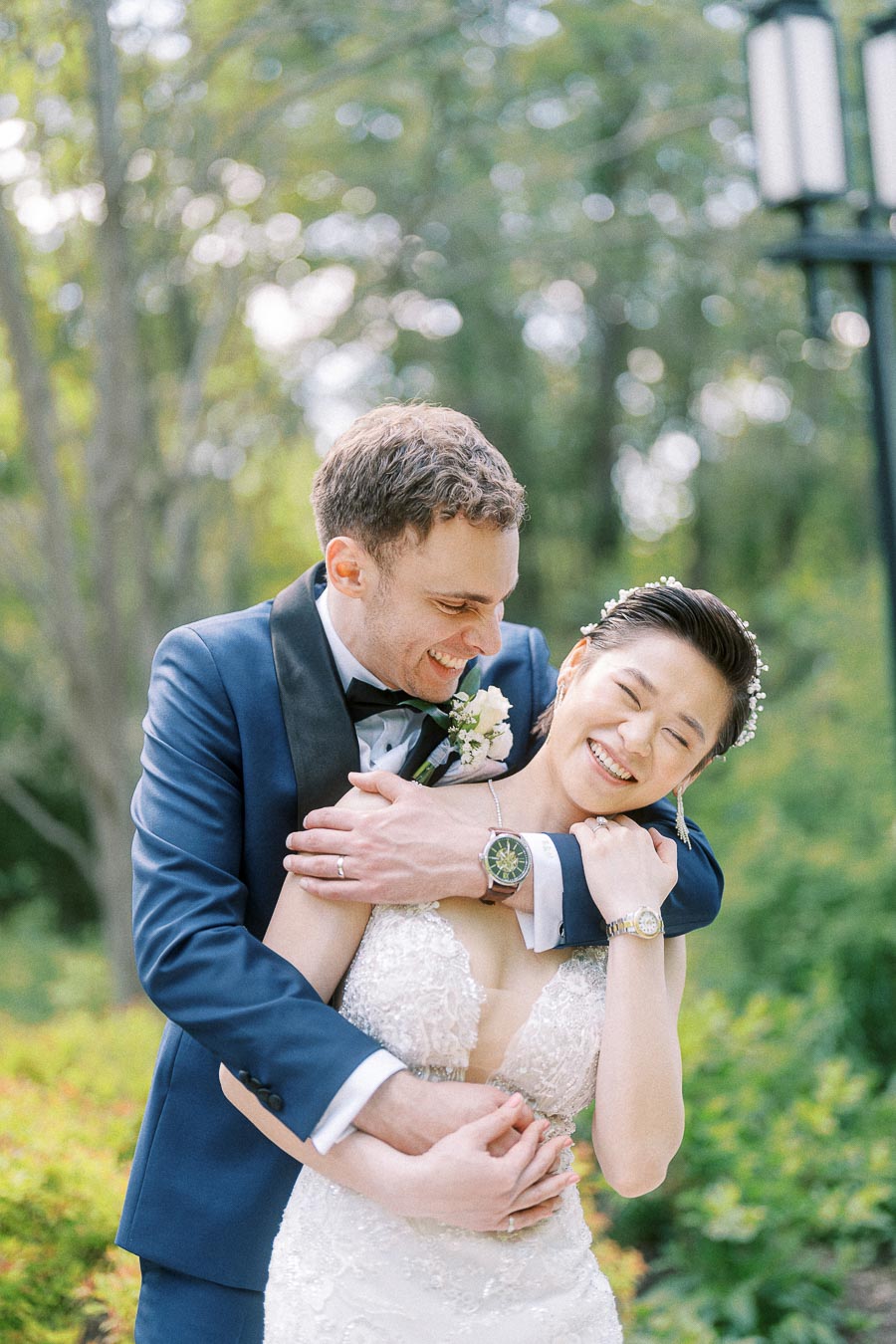 Groom in a navy tuxedo hugs the smiling bride from behind in a green garden setting.