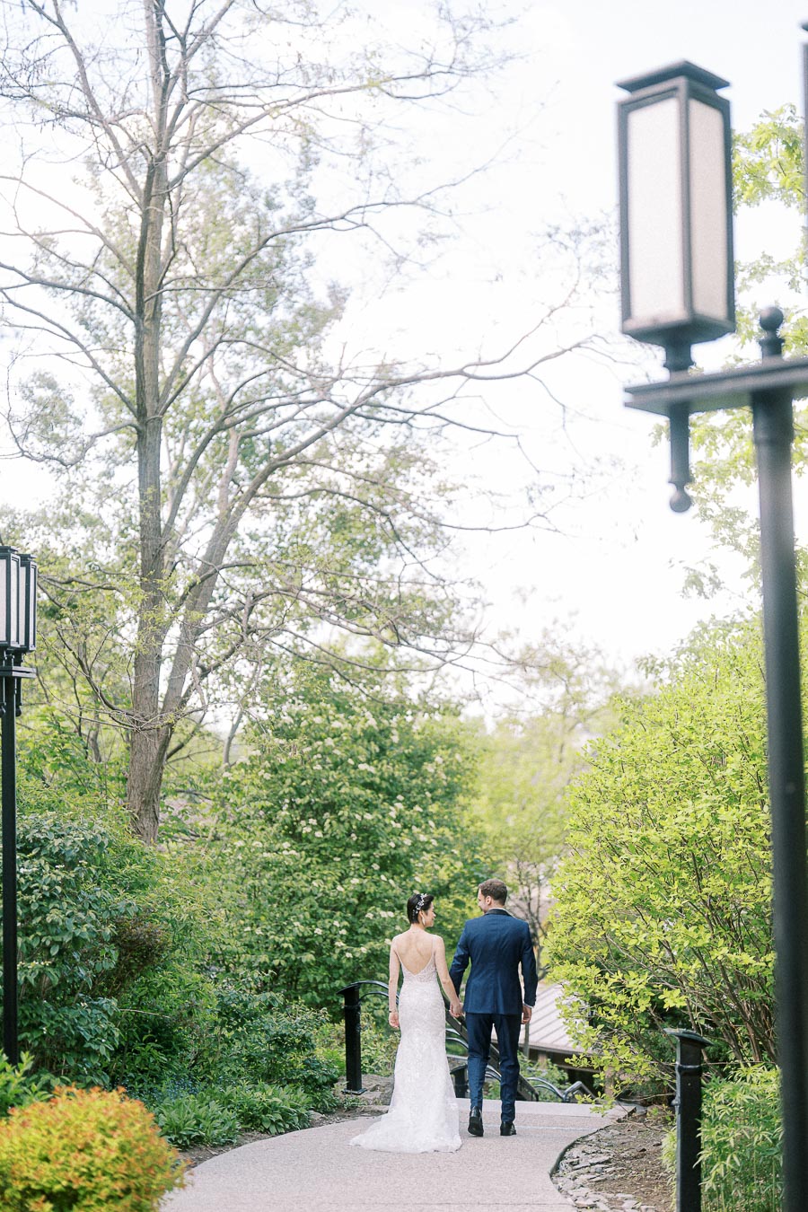 Bride and groom walk hand in hand along a garden path with lanterns and spring greenery at Ancaster Mill.