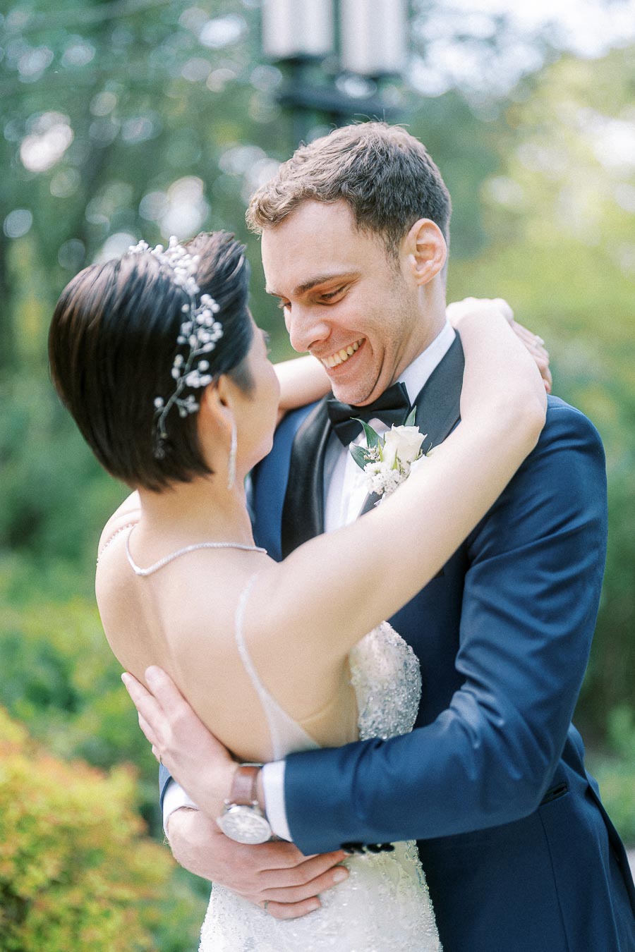 Groom smiles while embracing the bride in a beaded gown and floral hairpiece outdoors.