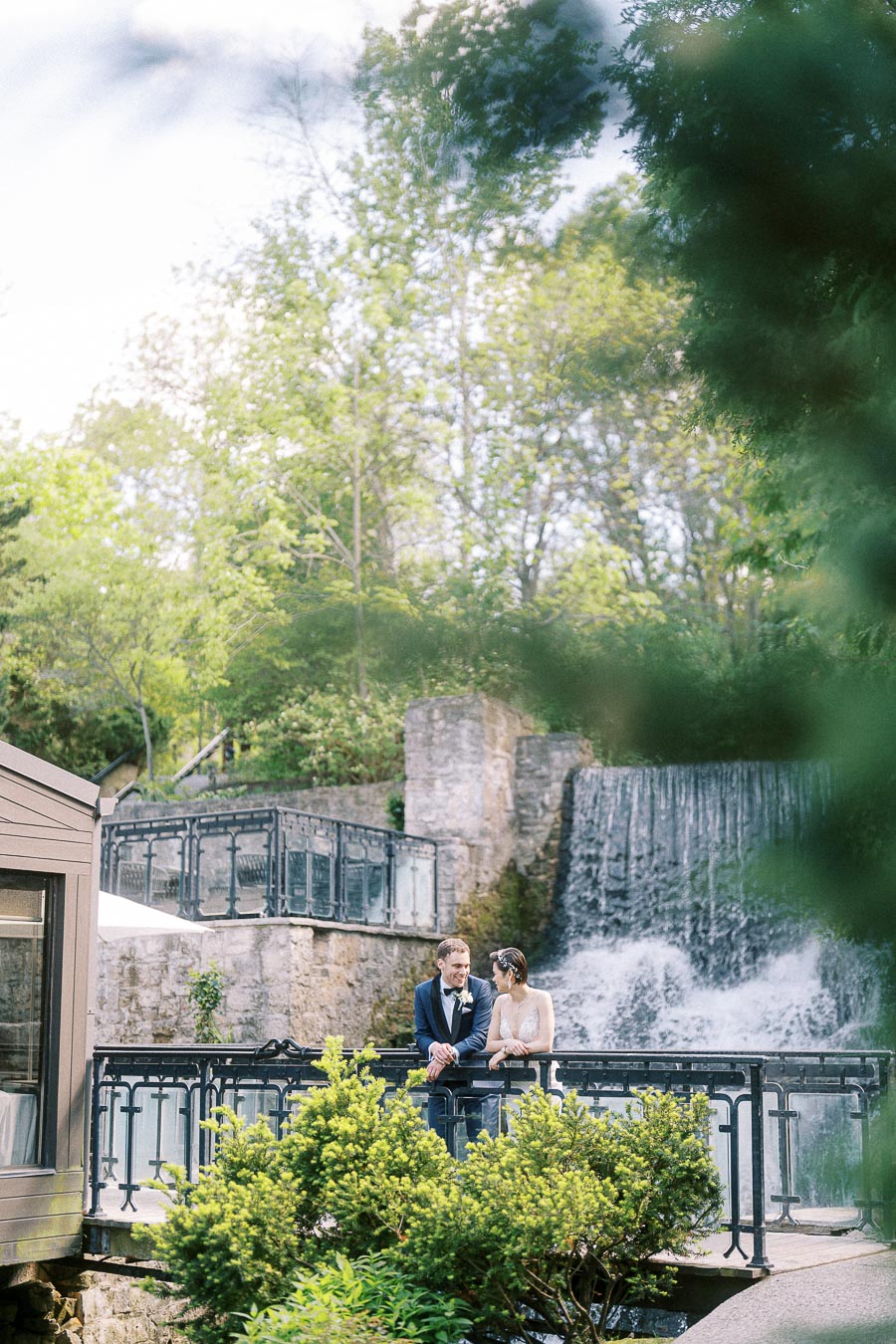 Bride and groom standing on a railing-lined bridge beside the waterfall at Ancaster Mill.