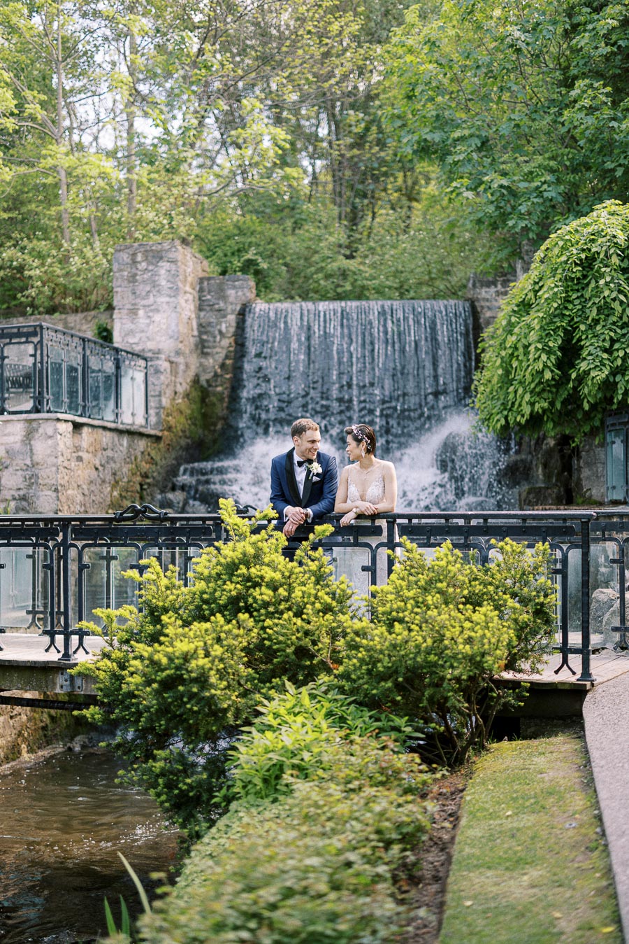 Miffy and Rawle stand together on a bridge in wedding attire with a waterfall and greenery behind them.