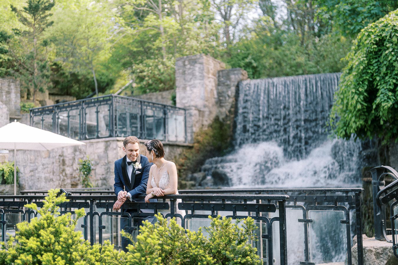 Bride and groom lean on a black railing beside the Ancaster Mill waterfall surrounded by spring greenery.