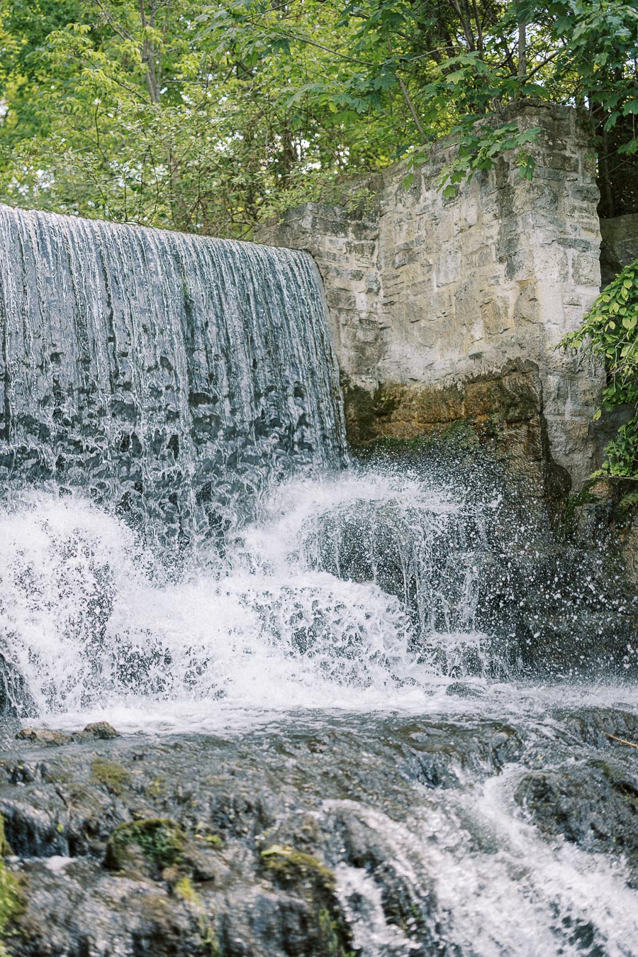 Water cascades over a stone mill waterfall surrounded by green trees and rock walls.