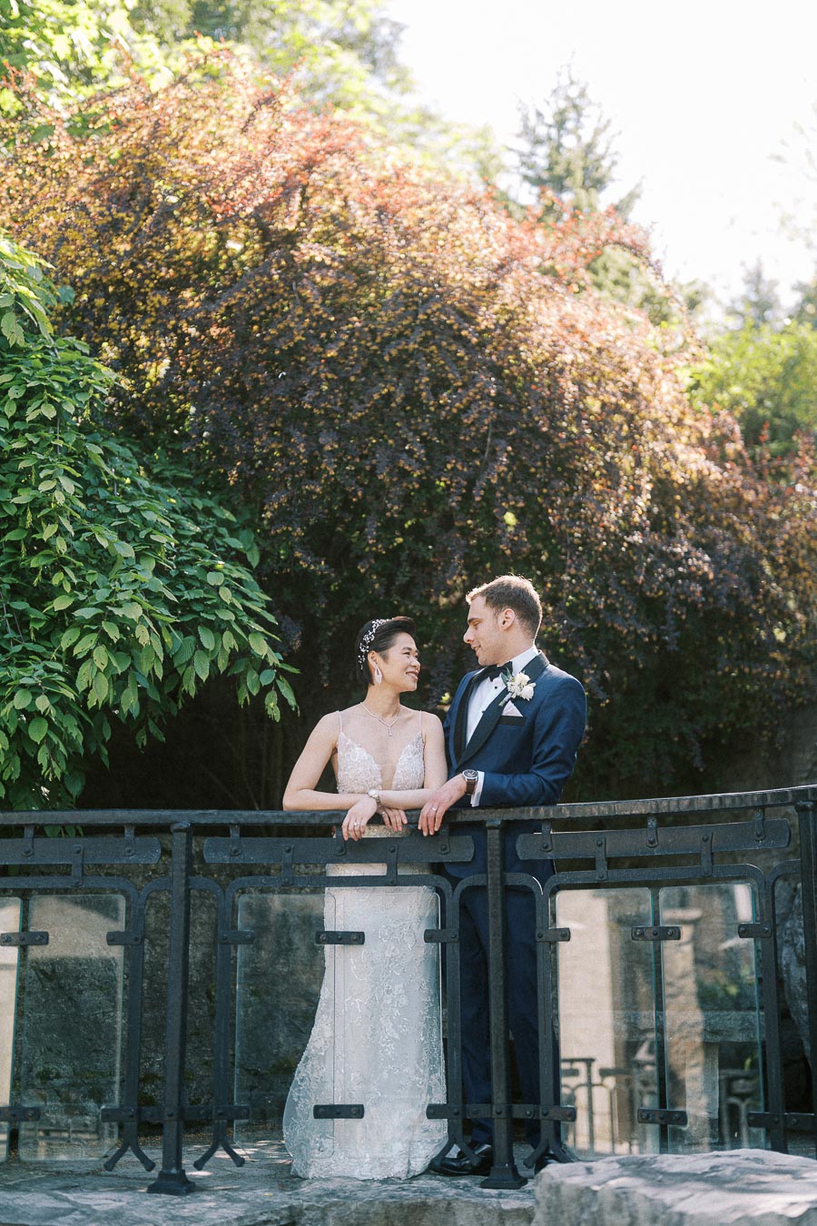 Bride and groom lean on a glass bridge railing, smiling at each other amid lush greenery at Ancaster Mill.