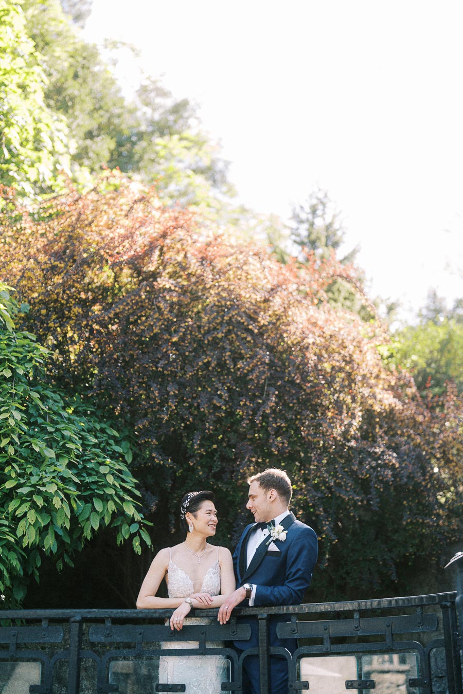 Miffy and Rawle smile at each other while leaning on a garden bridge railing in wedding attire.