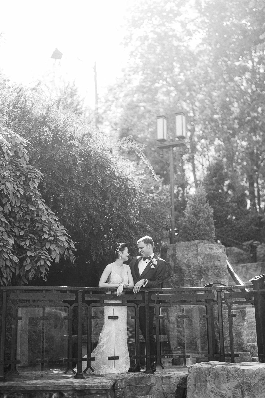 Bride and groom in wedding attire leaning on a bridge railing in a wooded garden setting.