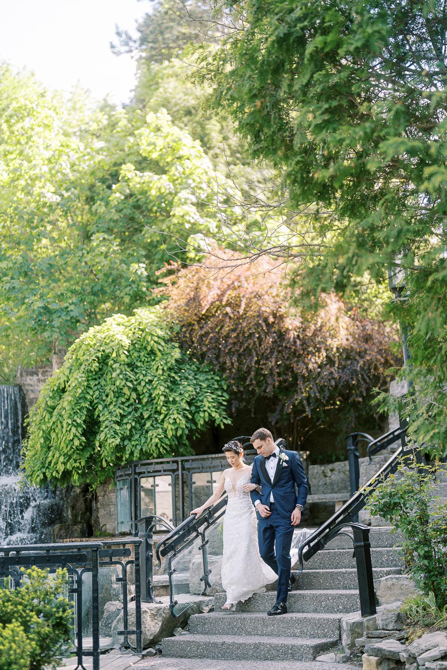 Bride and groom walk down stone steps beside a waterfall and greenery at Ancaster Mill.
