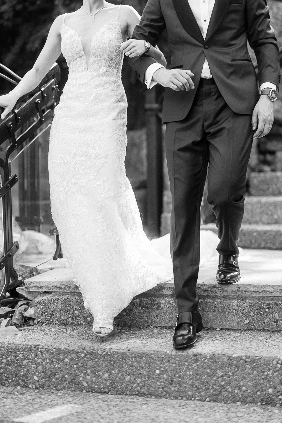Bride in a lace gown and groom in a tuxedo walk arm in arm down stone steps beside a railing.