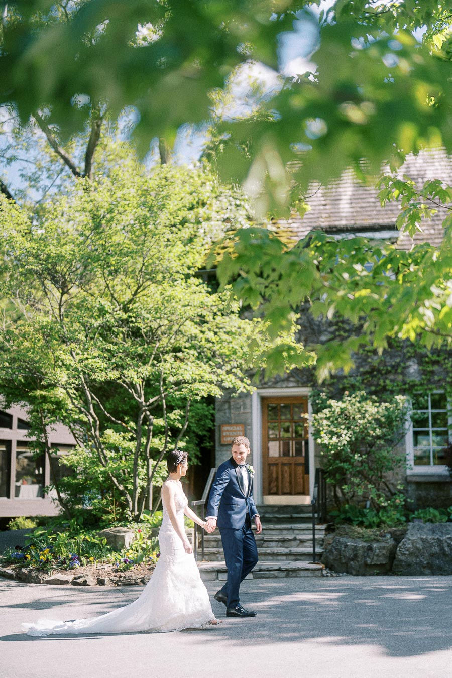 Bride and groom holding hands outside a stone building surrounded by leafy trees at Ancaster Mill.