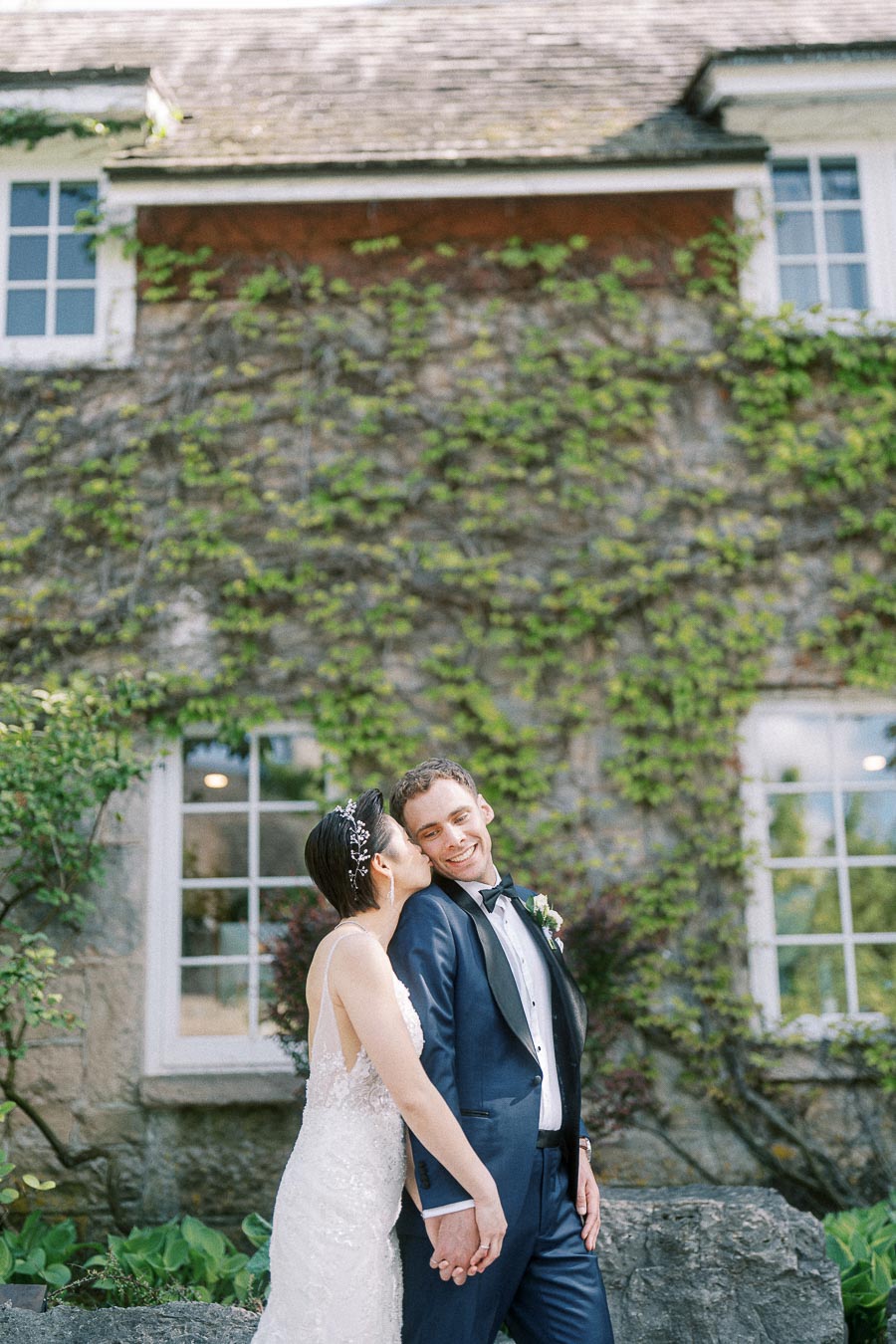 Bride kisses smiling groom while holding hands outside an ivy-covered stone building at Ancaster Mill.
