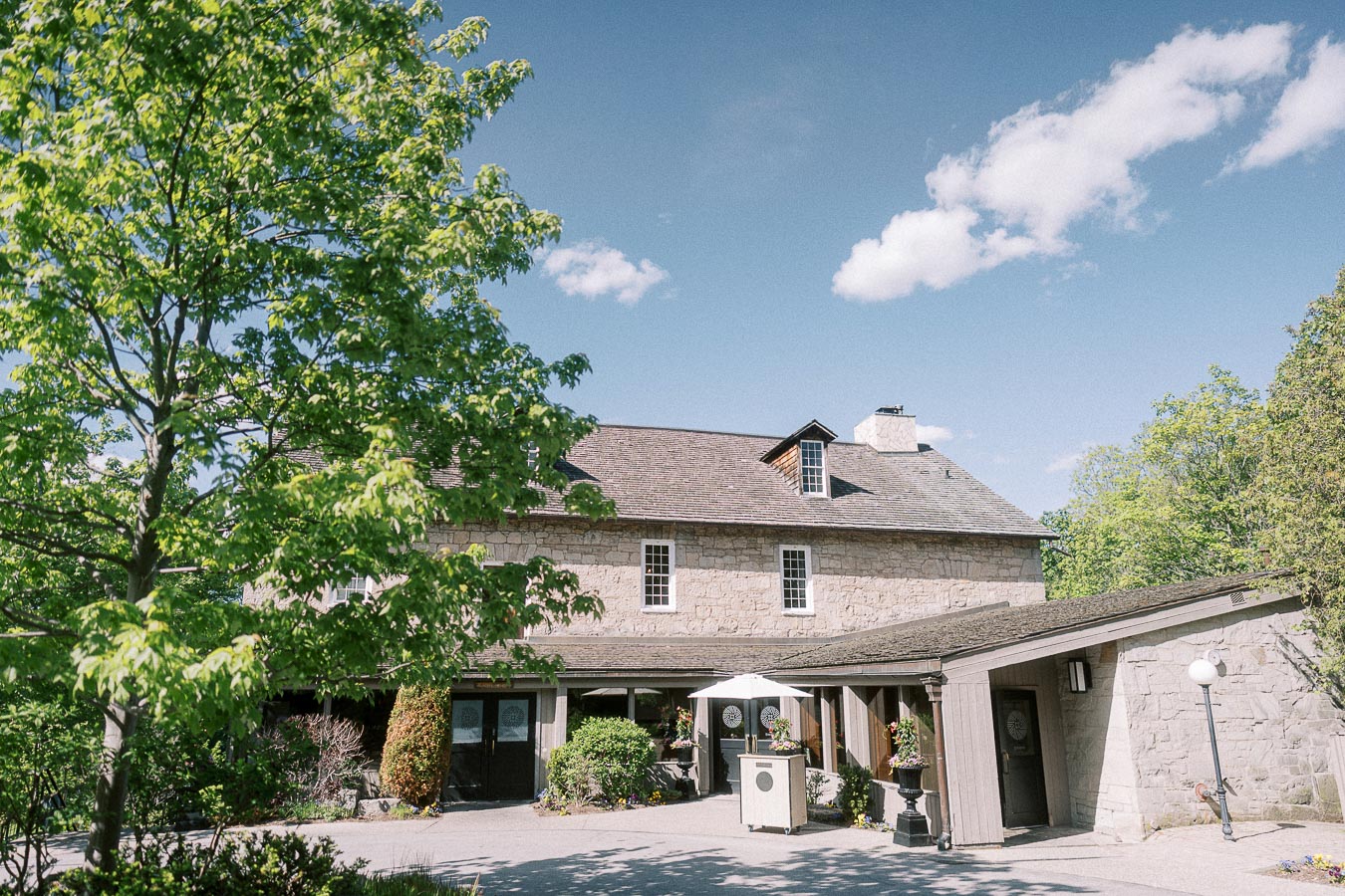 Stone building at Ancaster Mill with leafy trees, patio umbrellas, and a bright blue sky