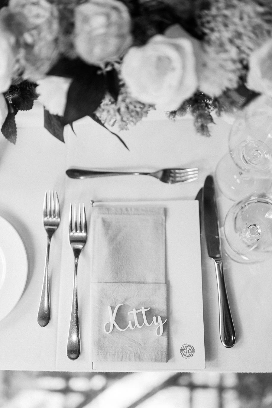 Black-and-white place setting with a Kitty name card, folded napkin, cutlery, glasses, and floral centerpiece.