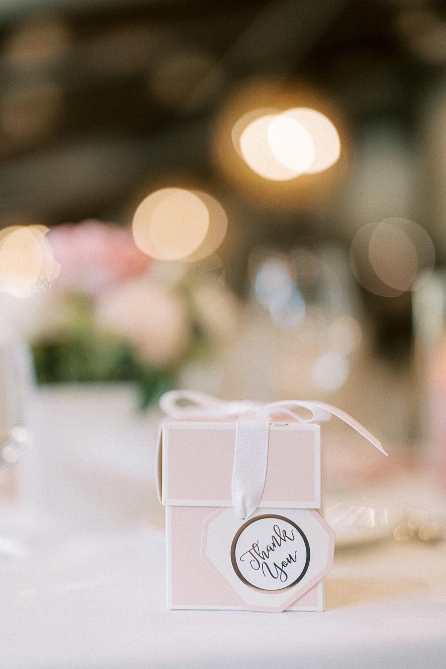 Pink wedding favor box with a white ribbon and thank you tag on a softly lit reception table.