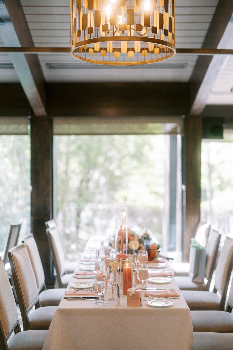 Long wedding reception table with coral candles, place settings, floral accents, and a chandelier overhead