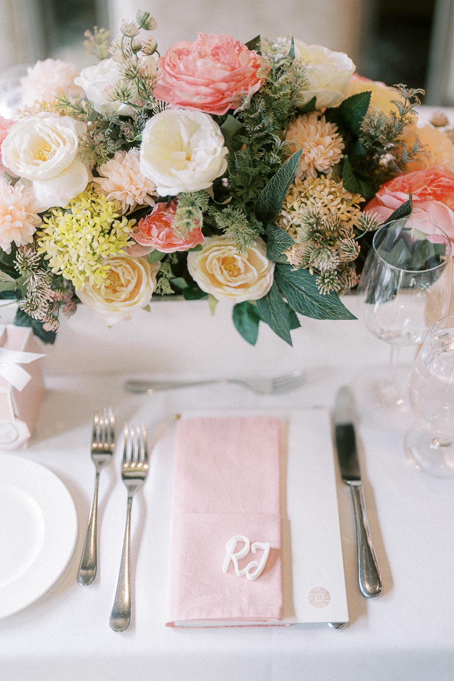 Pastel floral centerpiece above a place setting with pink napkin and RJ initials at a wedding reception