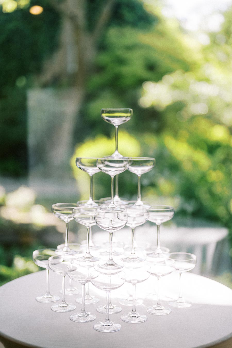 Tiered champagne coupe tower on a white table outdoors with soft greenery and waterfall blur behind it