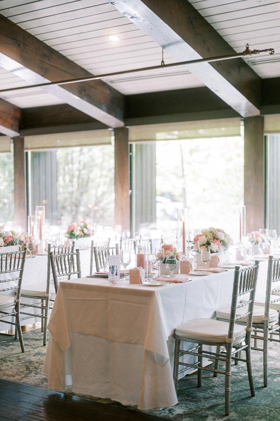 Reception tables with white linens, silver chairs, pink florals and tall candles in a bright timber-beamed room.