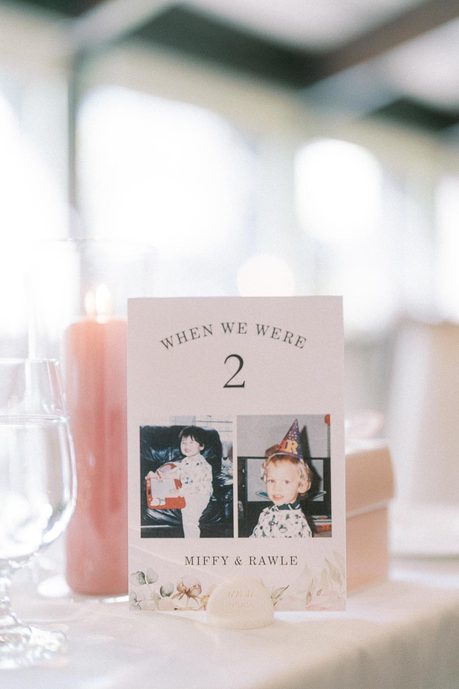 Reception table number card for Miffy and Rawle with childhood photos at age two beside a pink candle.