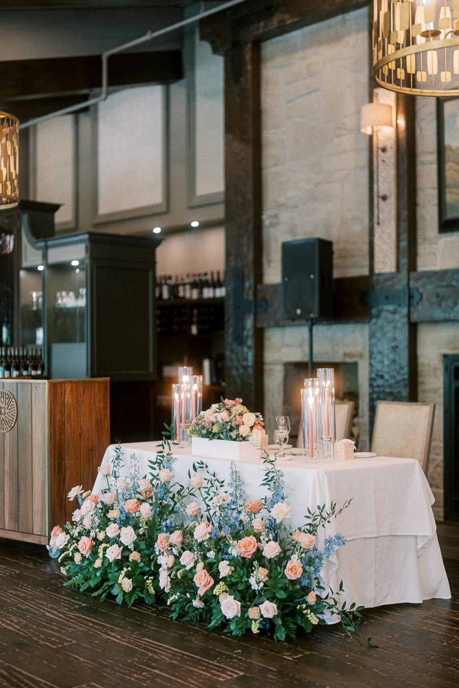 Head table with pastel floral arrangements and tall candles inside a rustic wedding reception room.