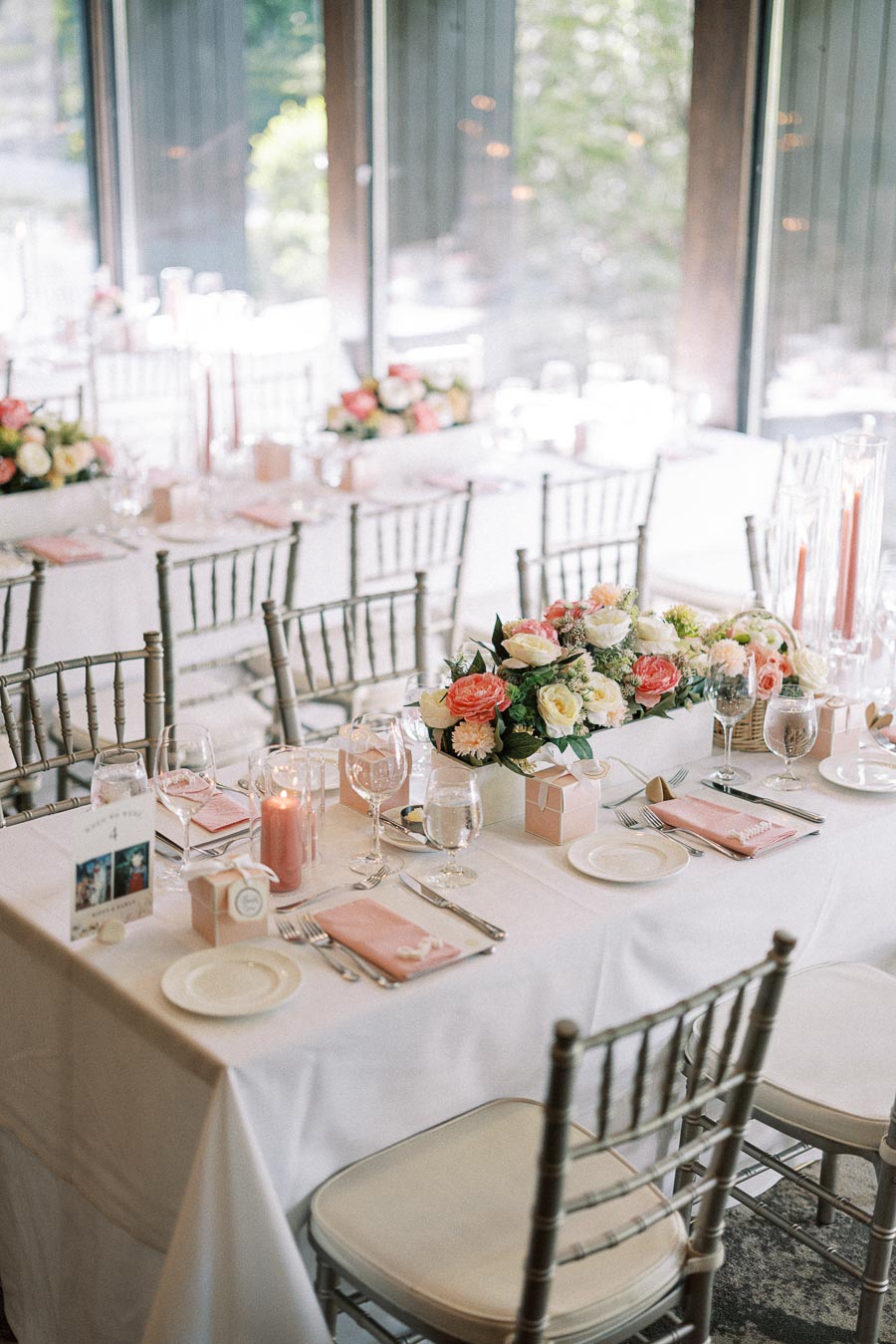 Elegant wedding reception tables with peach napkins, floral centerpieces, candles, glassware, and silver chairs by tall windows.