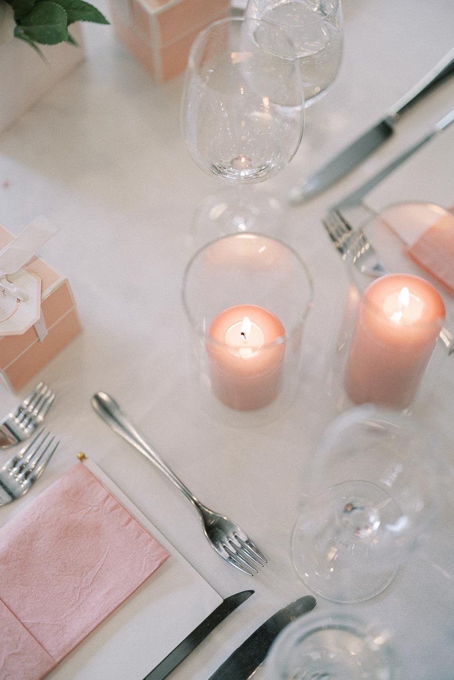 Reception table setting with pink candles, folded napkins, wine glasses, cutlery, and favor boxes on white linen.
