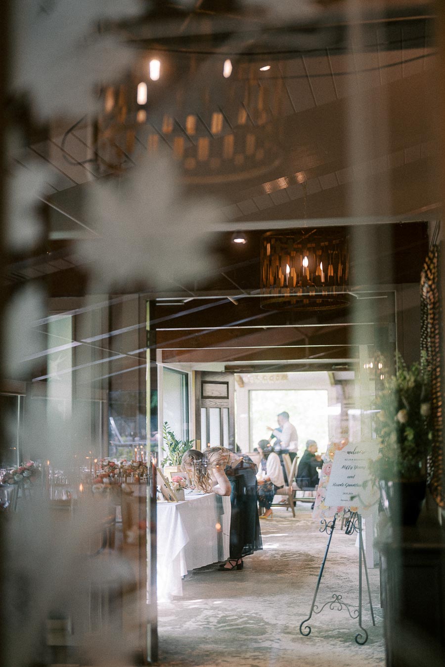 Guests gather at a wedding reception entrance with a seating sign, tables, and warm chandeliers visible through glass.
