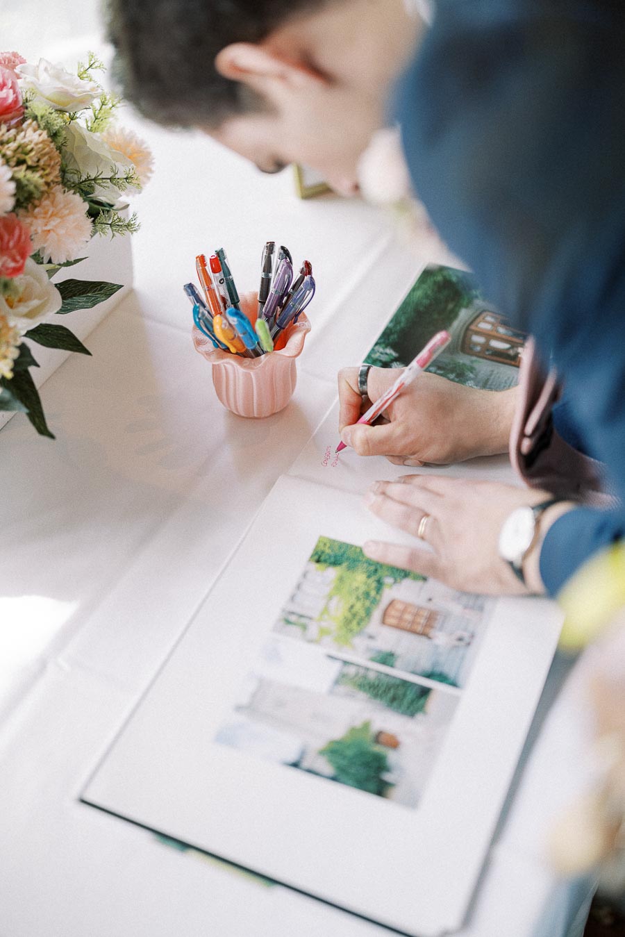 Guest signs a wedding guest book beside colorful pens and pastel flowers on a white table.