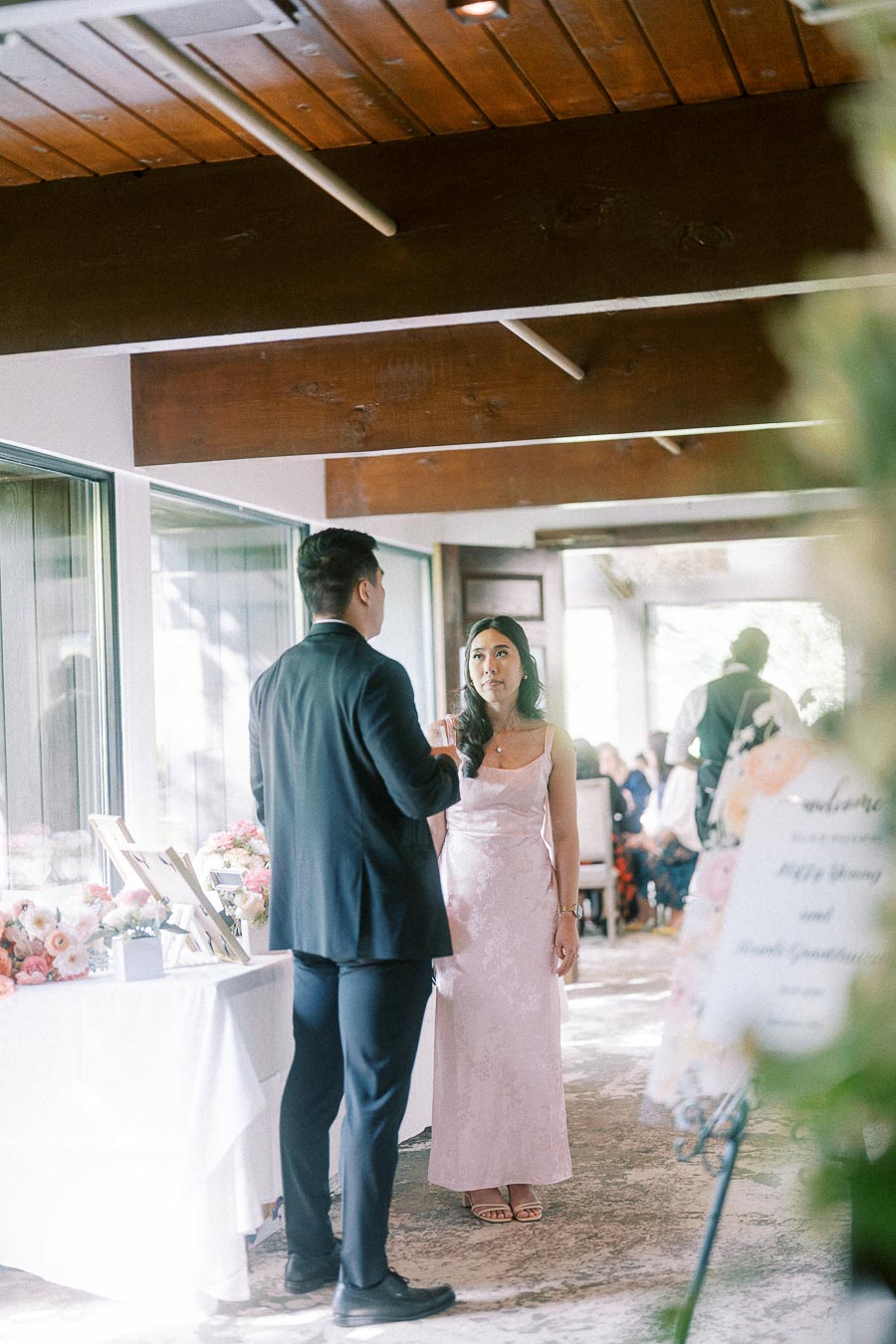 Man in a dark suit speaks with a woman in a pink dress beside floral displays at an indoor wedding reception.