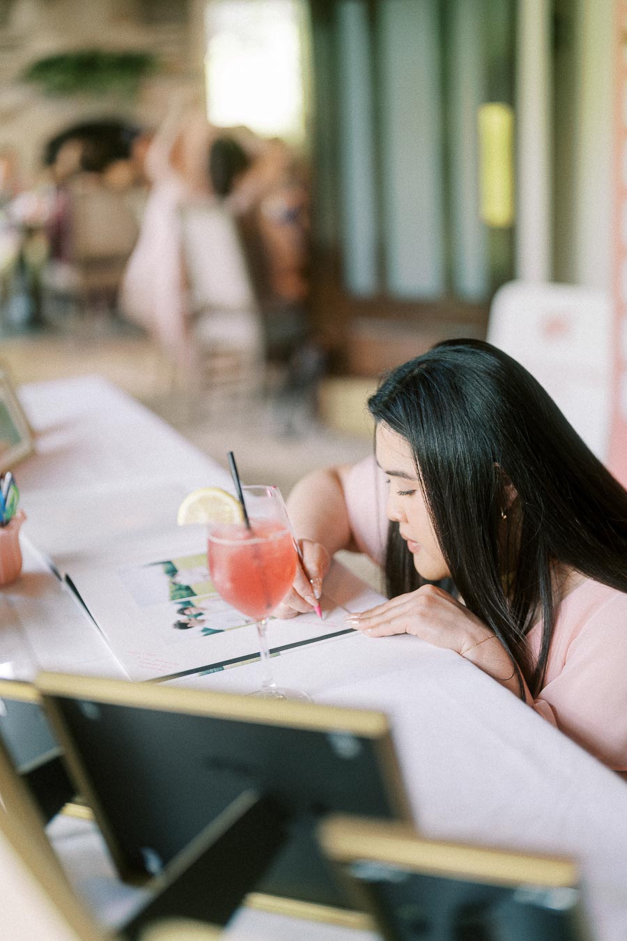 Woman in a pink dress writes in a wedding guest book beside a pink drink on a reception table.
