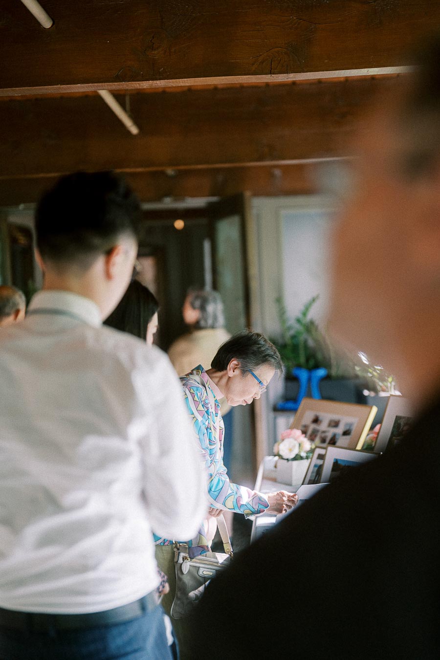 Guest in a patterned shirt signs beside a framed photo display while others gather indoors at the wedding.