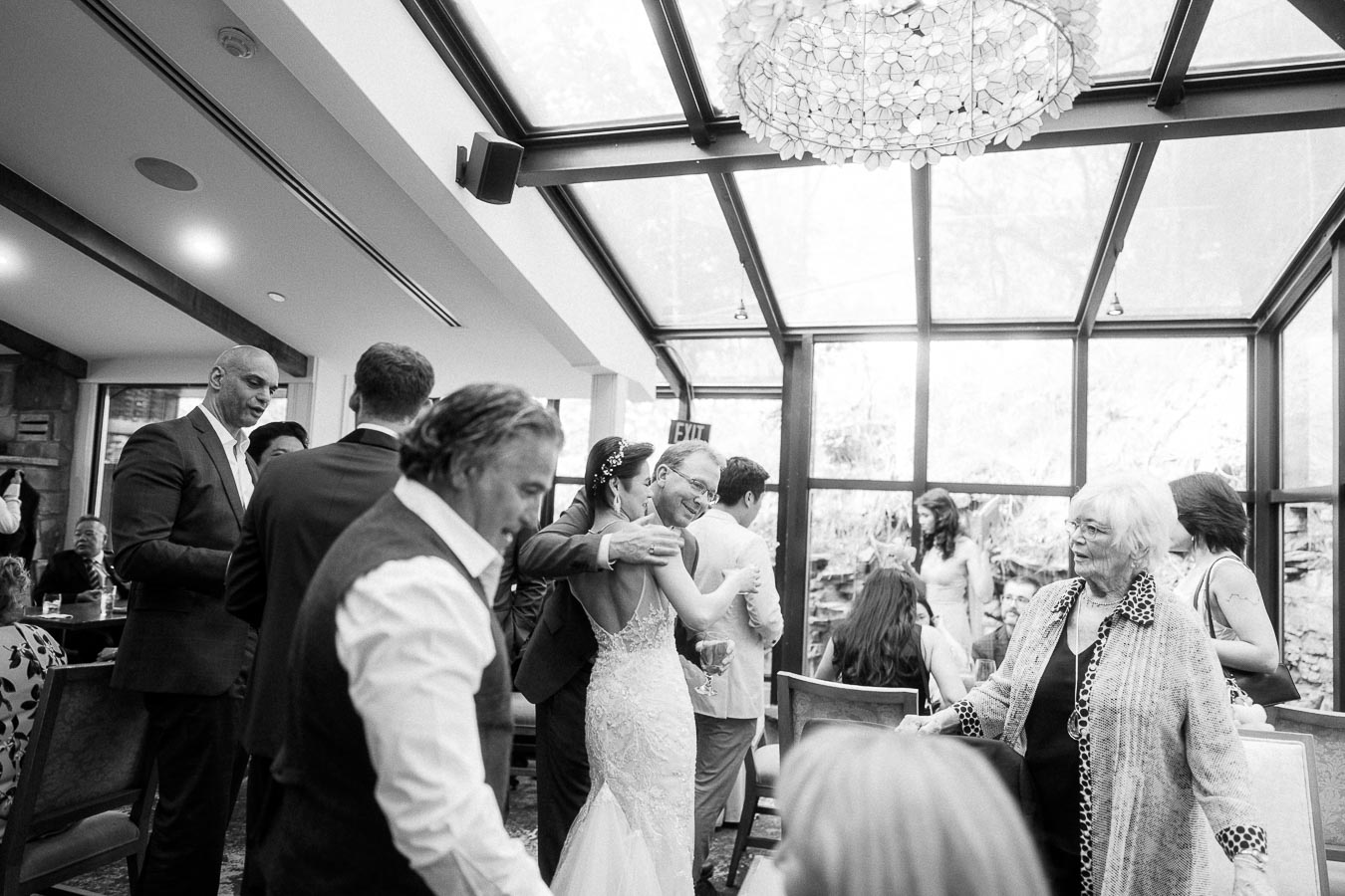 Bride embraces an older guest in a bright glass-walled reception room as wedding guests mingle nearby.